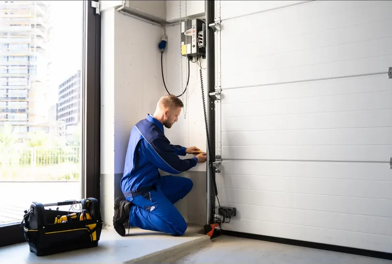Technician kneeling beside a white garage door working on the cable and track along the side rail